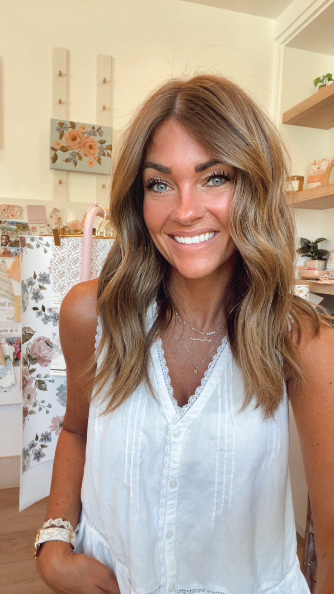Mindy Young with wavy light brown hair wearing a white sleeveless top, standing in a brightly lit boutique interior with shelves and floral decor in the background.