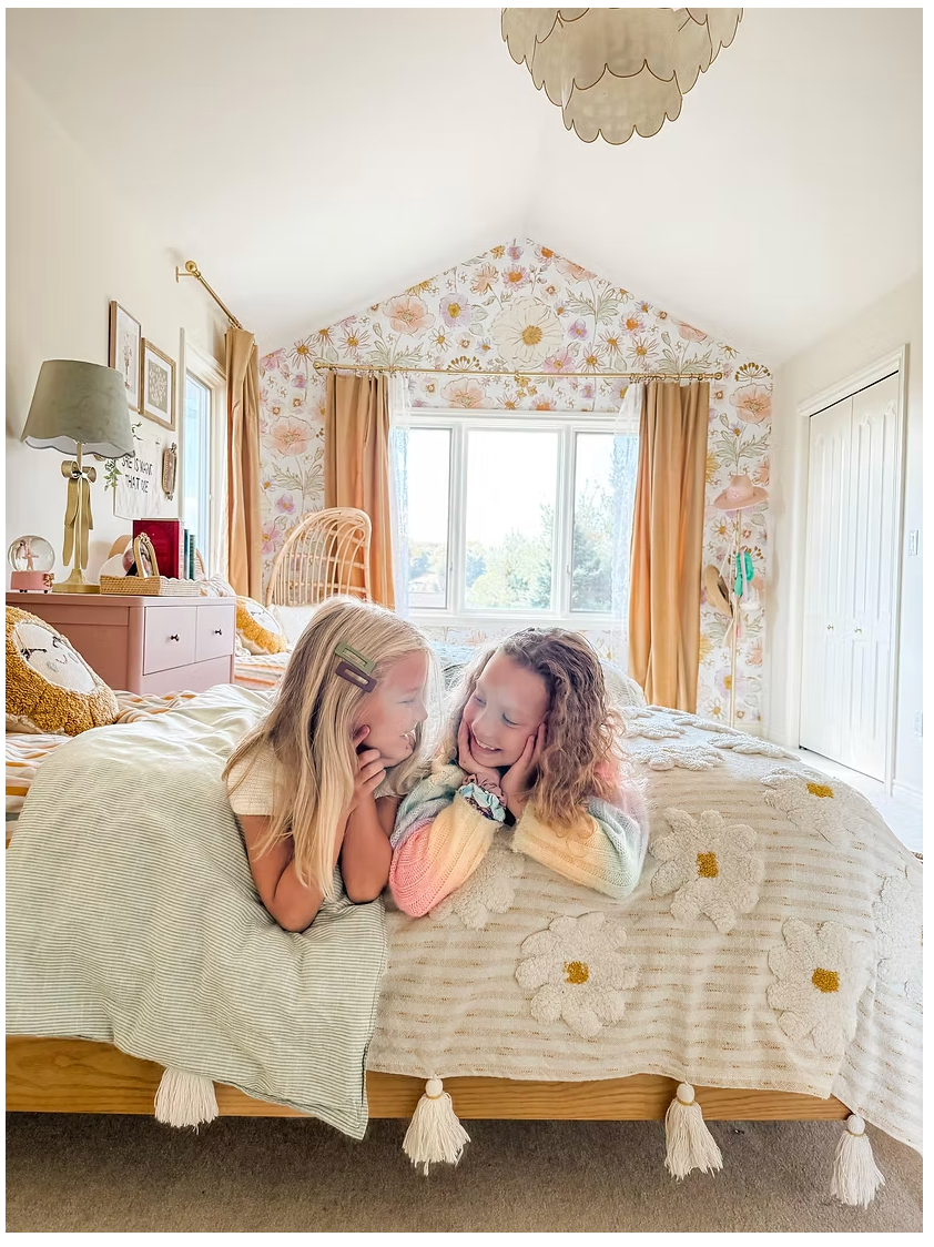 Two children playing on a bed in a bedroom with floral curtains and a lamp.